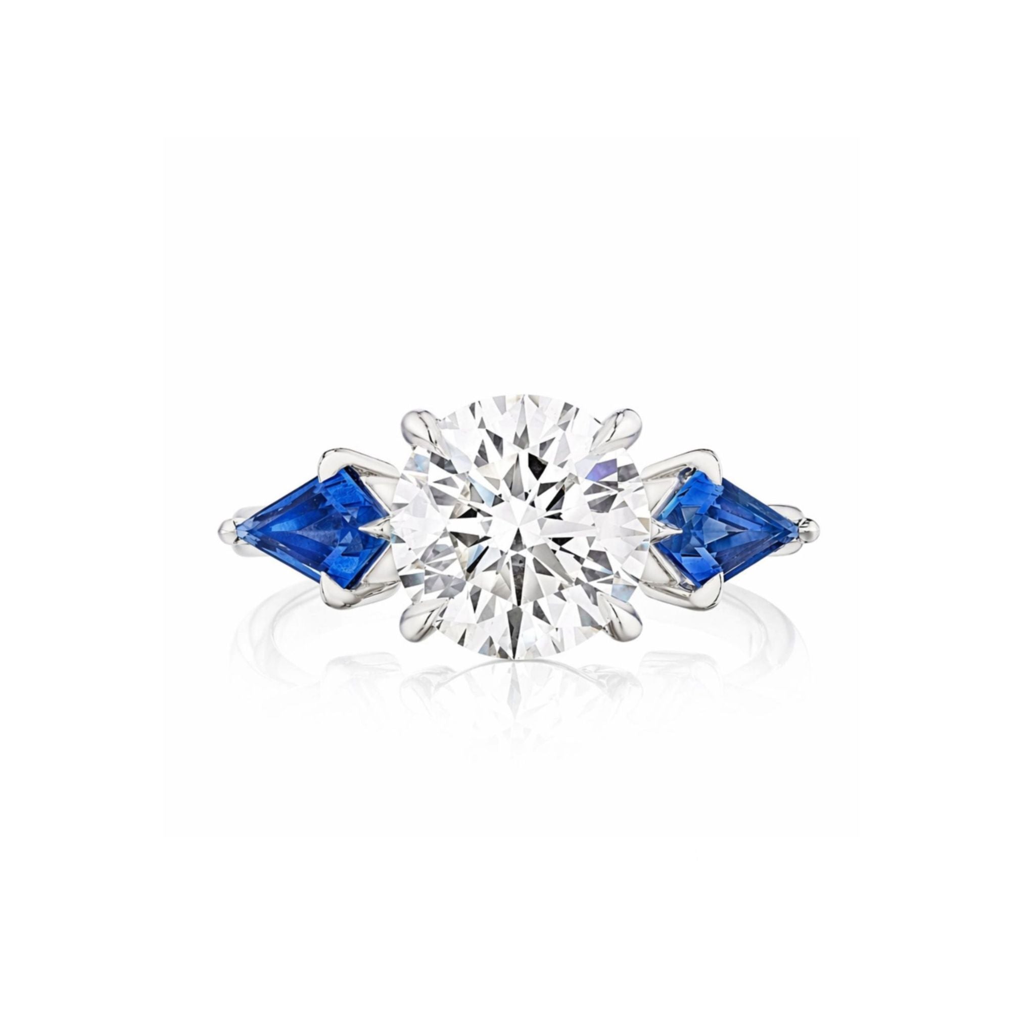 Introducing the Round Diamond Engagement Ring in White Gold with Kite Shape Blue Sapphire Side Stones – a breathtaking design featuring a brilliant round center diamond, accented by uniquely shaped kite blue sapphires on either side and set gracefully on a sleek white gold band.