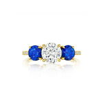 Introducing the Round Diamond Engagement Ring with Round Sapphire Side Stones—crafted in exquisite 18k yellow gold, this stunning ring showcases a large round brilliant diamond at its center, complemented by vibrant blue round sapphire side stones and sparkling accent diamonds set along the band.