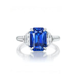 The Emerald Cut Sapphire Engagement Ring with Half Moon Diamond Side Stones showcases a stunning emerald-cut blue sapphire at its center, flanked on each side by sparkling half moon diamond accents, all set against a plain white background.
