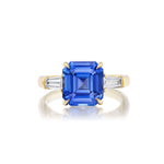 The Asscher Cut Sapphire Engagement Ring with Tapered Baguette Diamond Side Stones showcases a bold Asscher cut sapphire set in luxurious 18k yellow gold, elegantly accented by pairs of tapered baguette diamonds on each side, photographed against a pristine white background.