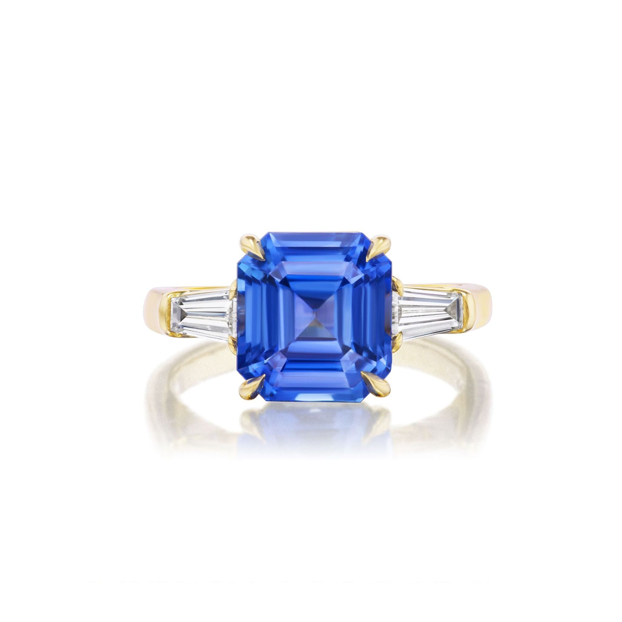 The Asscher Cut Sapphire Engagement Ring with Tapered Baguette Diamond Side Stones showcases a bold Asscher cut sapphire set in luxurious 18k yellow gold, elegantly accented by pairs of tapered baguette diamonds on each side, photographed against a pristine white background.
