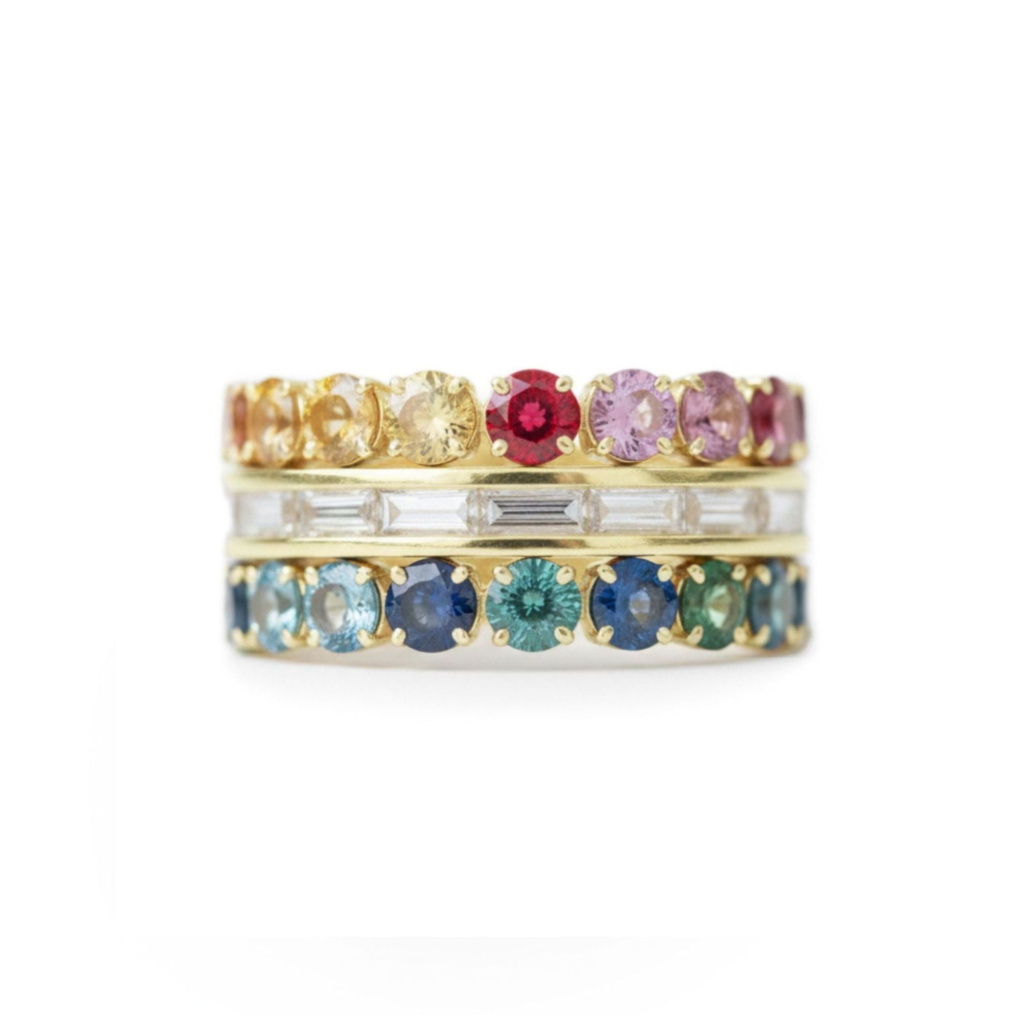 Three gold bands are stacked, featuring one with multicolor round sapphires and another with channel set baguette diamonds, all set in yellow gold.