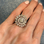 A hand with light pink nails wears the Spiral Ring in White Gold with Round Brilliant and Baguette Diamonds, featuring an ornate design with a large central diamond surrounded by sparkling baguette and round brilliant diamonds, displayed against a denim background.
