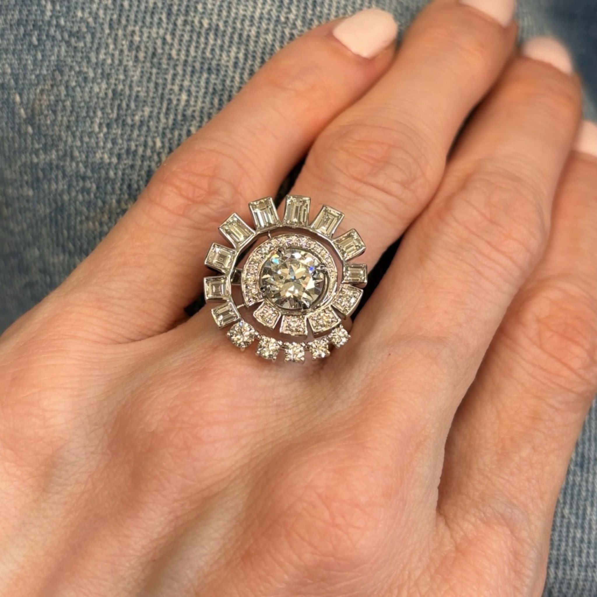 A hand with light pink nails wears the Spiral Ring in White Gold with Round Brilliant and Baguette Diamonds, featuring an ornate design with a large central diamond surrounded by sparkling baguette and round brilliant diamonds, displayed against a denim background.