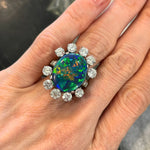 A hand wears the Sunburst Oval Opal Ring, showcasing a vibrant oval opal encircled by a halo of sparkling round brilliant diamonds and pavé accents, all set in an elegant 18k white gold band.