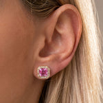 A close-up of a woman's ear showcases the Mosaic Studs in Rose Gold, featuring a striking pink asscher cut sapphire set in a geometric design and framed by glittering baguette diamonds. Her blonde hair partially falls over the ear, highlighting the intricate mosaic details that surround the radiant gemstone.