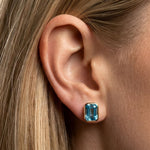 Close-up of a woman's ear adorned with the Bezel Set Emerald Cut Aquamarine Stud Earrings in Yellow Gold, her straight blonde hair tucked behind her ear.