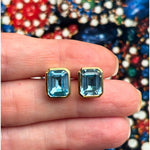 A hand displays the Bezel Set Emerald Cut Aquamarine Stud Earrings in Yellow Gold against a colorful, blurred background.