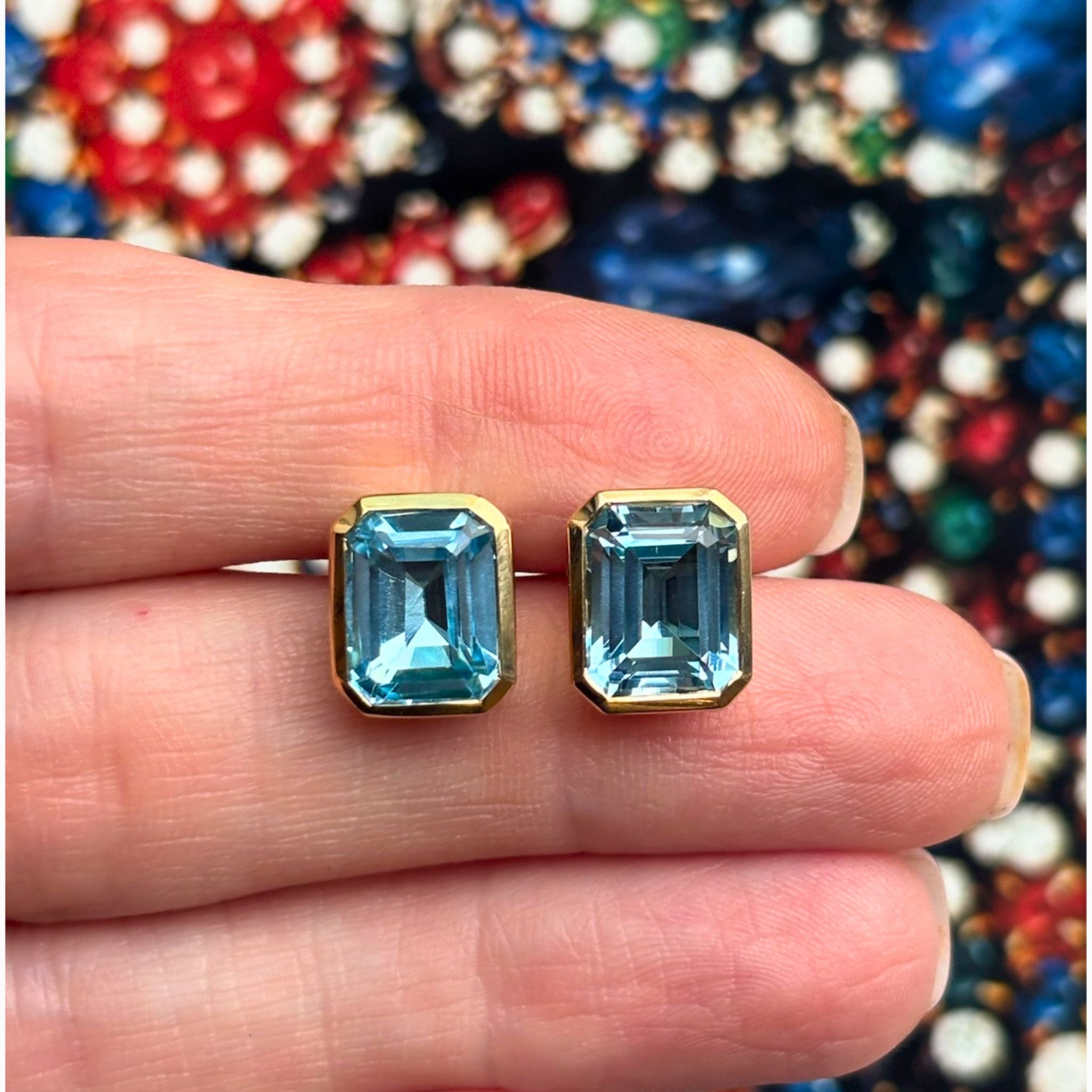 A hand displays the Bezel Set Emerald Cut Aquamarine Stud Earrings in Yellow Gold against a colorful, blurred background.