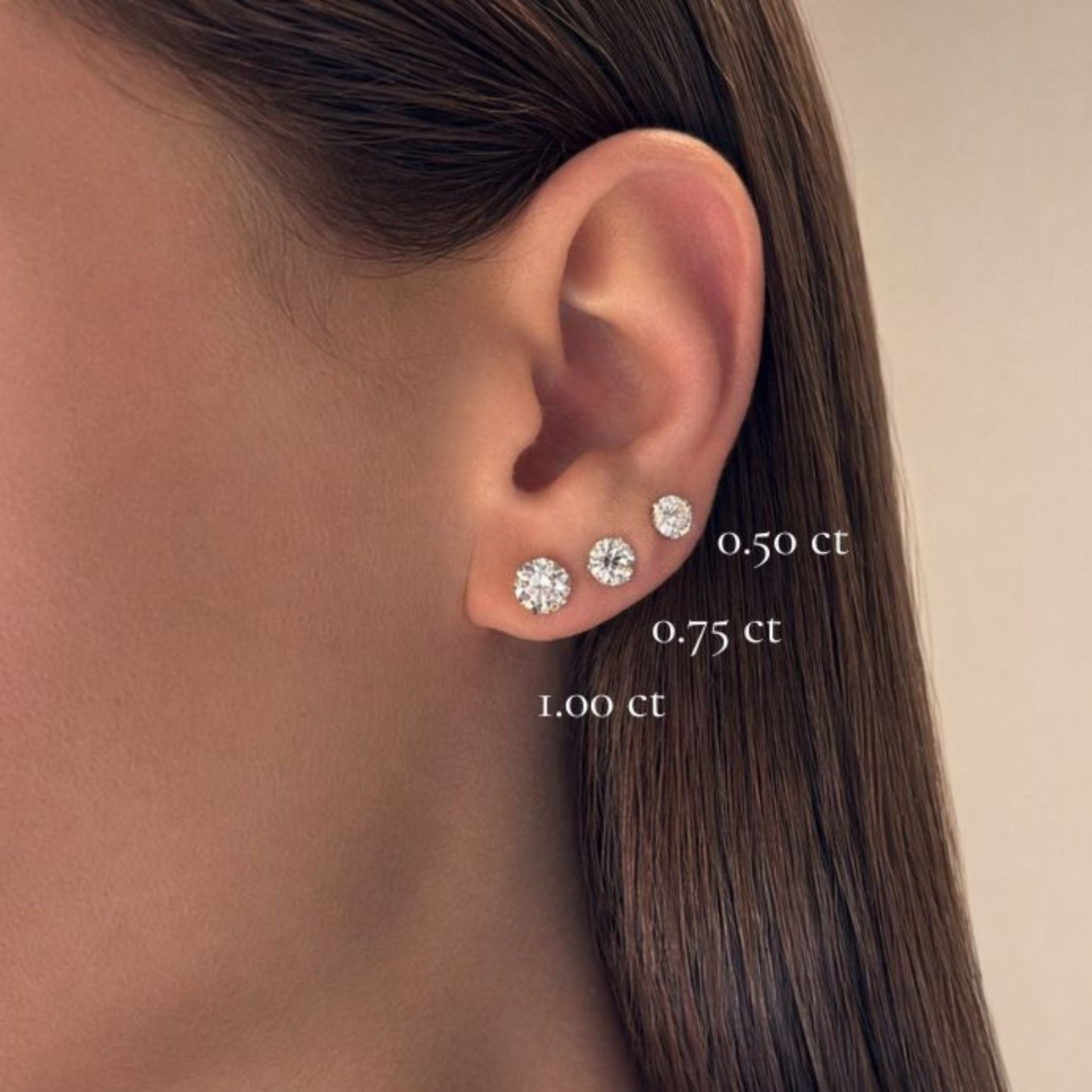 A close-up of a woman's ear wearing three Classic Round Diamond Studs in Martini Setting of different sizes—0.50 ct, 0.75 ct, and 1.00 ct—each crafted in luxurious 18k white gold and arranged from smallest to largest on the earlobe.