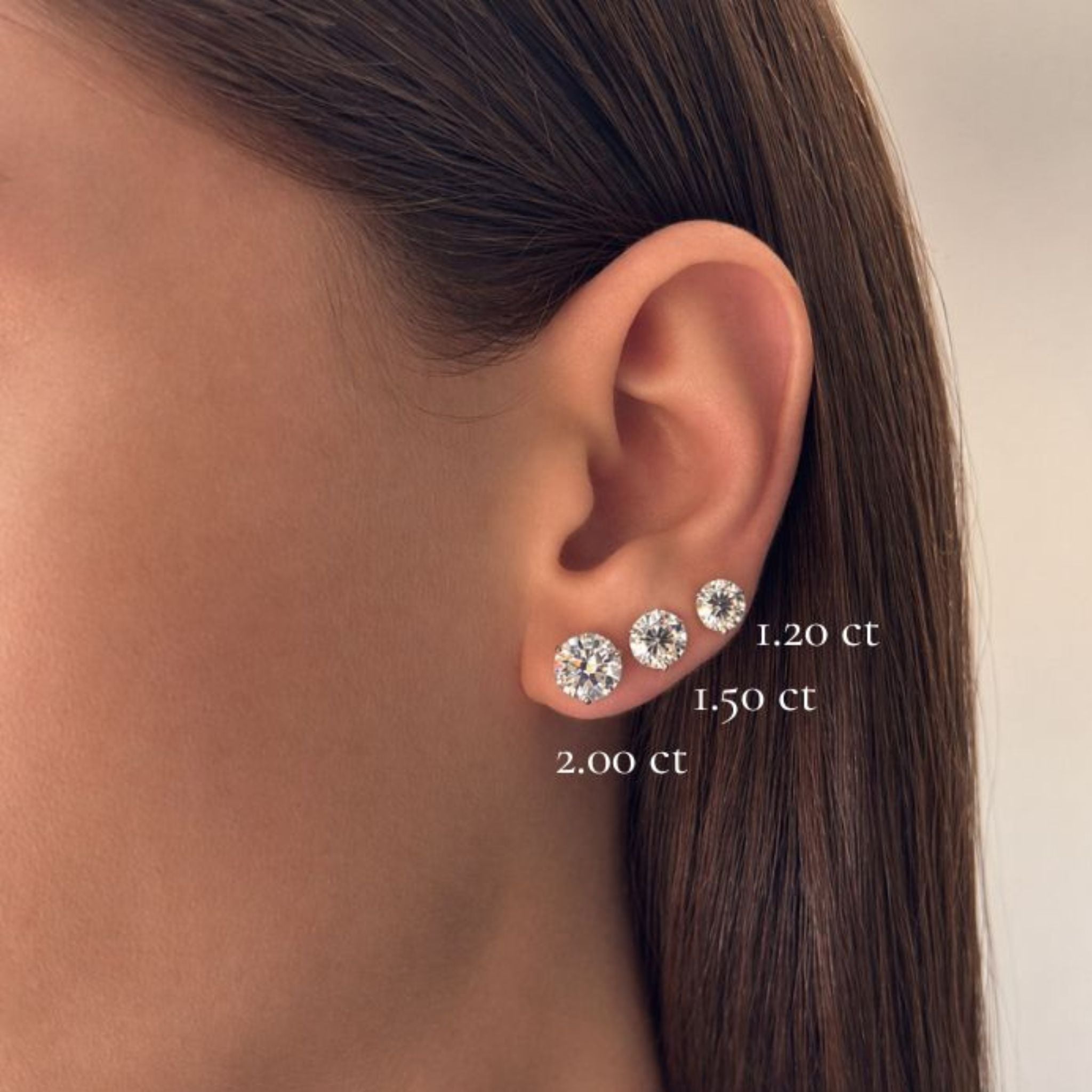 A close-up of an ear adorned with three Classic Round Diamond Studs in Martini Setting, featuring GIA Certified round brilliant diamonds in 18k white gold and graduated sizes labeled 1.20 ct, 1.50 ct, and 2.00 ct from top to bottom.