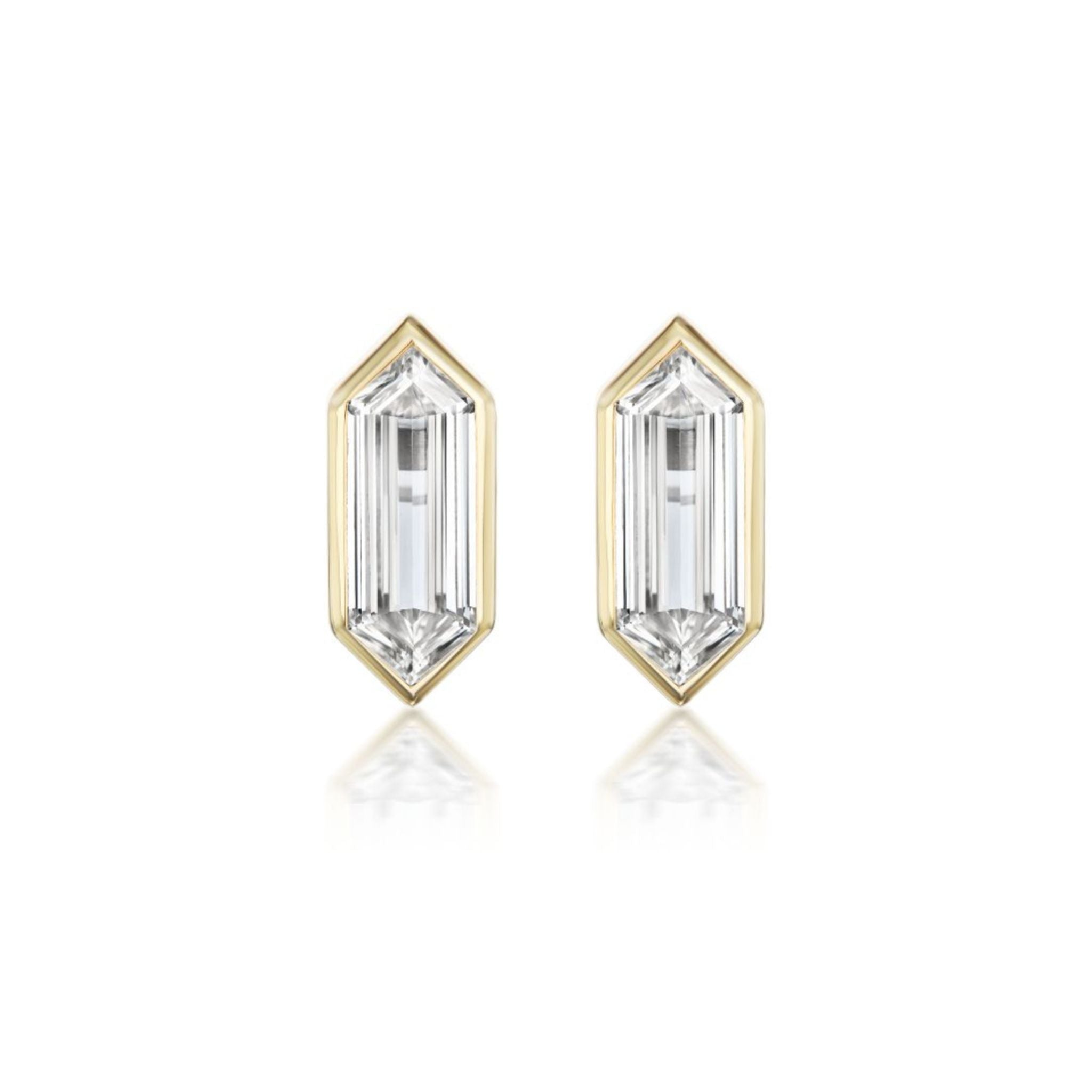 A pair of Bezel Set Studs in Yellow Gold with Elongated Hexagonal Diamonds is displayed against a white background, featuring emerald-cut clear gemstones set in hexagonal 18k yellow gold settings.