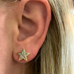 Close-up of a person's ear with straight blonde hair, wearing the Bezel Set Kite Diamond Star Studs in Yellow Gold—gold star-shaped earrings set with sparkling, clear gemstones that evoke the brilliance of a kite diamond.