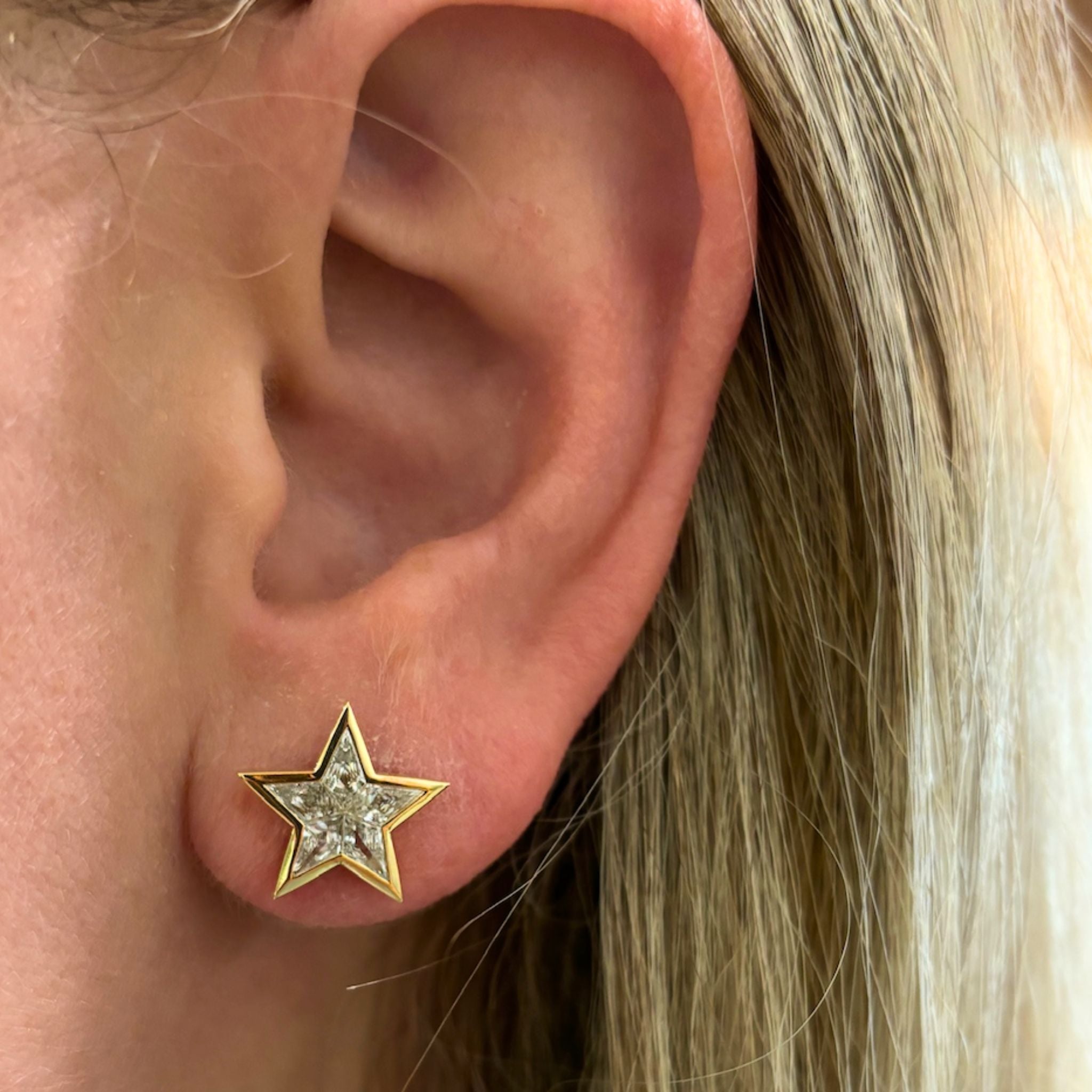 Close-up of a person's ear with straight blonde hair, wearing the Bezel Set Kite Diamond Star Studs in Yellow Gold—gold star-shaped earrings set with sparkling, clear gemstones that evoke the brilliance of a kite diamond.