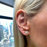 A close-up of a person's ear wearing the Diamond Star Studs in Yellow Gold—delicate star-shaped earrings set with small G-H color diamonds and crafted from 18k yellow gold—with a cityscape visible through a window in the background.