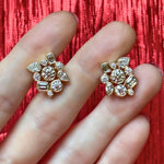Two fingers holding the Bezel Set Mixed Shape Diamond Cluster Earrings in yellow gold, featuring a cluster of round, marquise, and baguette-cut clear gemstones, set against a red textured background.
