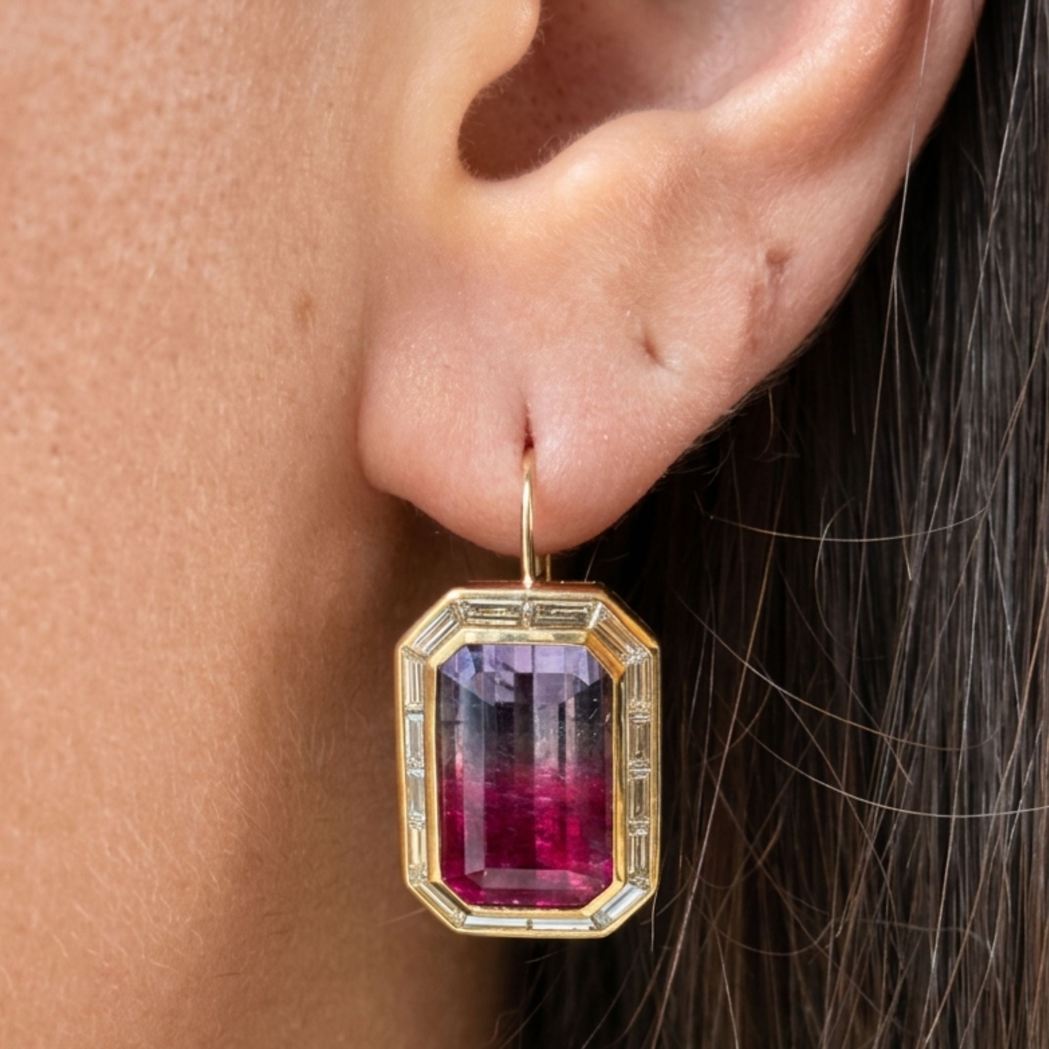 A close-up of an ear wearing the Watermelon Tourmaline Drop Earring with Baguette Diamond Halo in Yellow Gold, showcasing a vibrant rectangular gemstone framed in 18k yellow gold and accented by sparkling baguette diamonds. The gemstone displays pink and purple hues reminiscent of watermelon tourmalines, and the elegant earring hangs gracefully from a delicate gold hook.