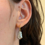A close-up of a person's ear with wavy brown hair, wearing the Collet Set Shield Shape Diamond Drop Earring, crafted from black rhodium and 18k yellow gold and featuring a geometric, shield-shaped diamond pendant.