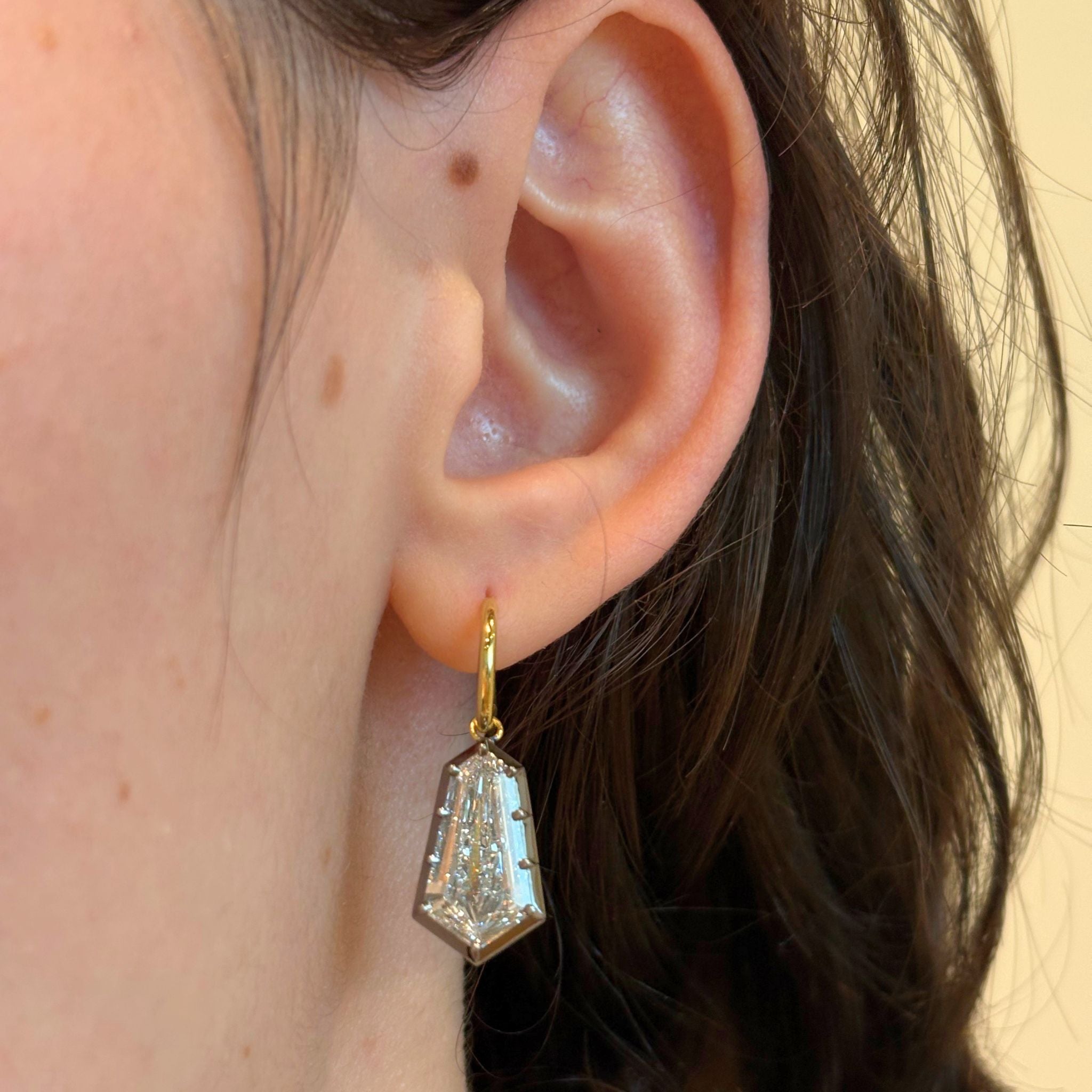 A close-up of a person's ear with wavy brown hair, wearing the Collet Set Shield Shape Diamond Drop Earring, crafted from black rhodium and 18k yellow gold and featuring a geometric, shield-shaped diamond pendant.