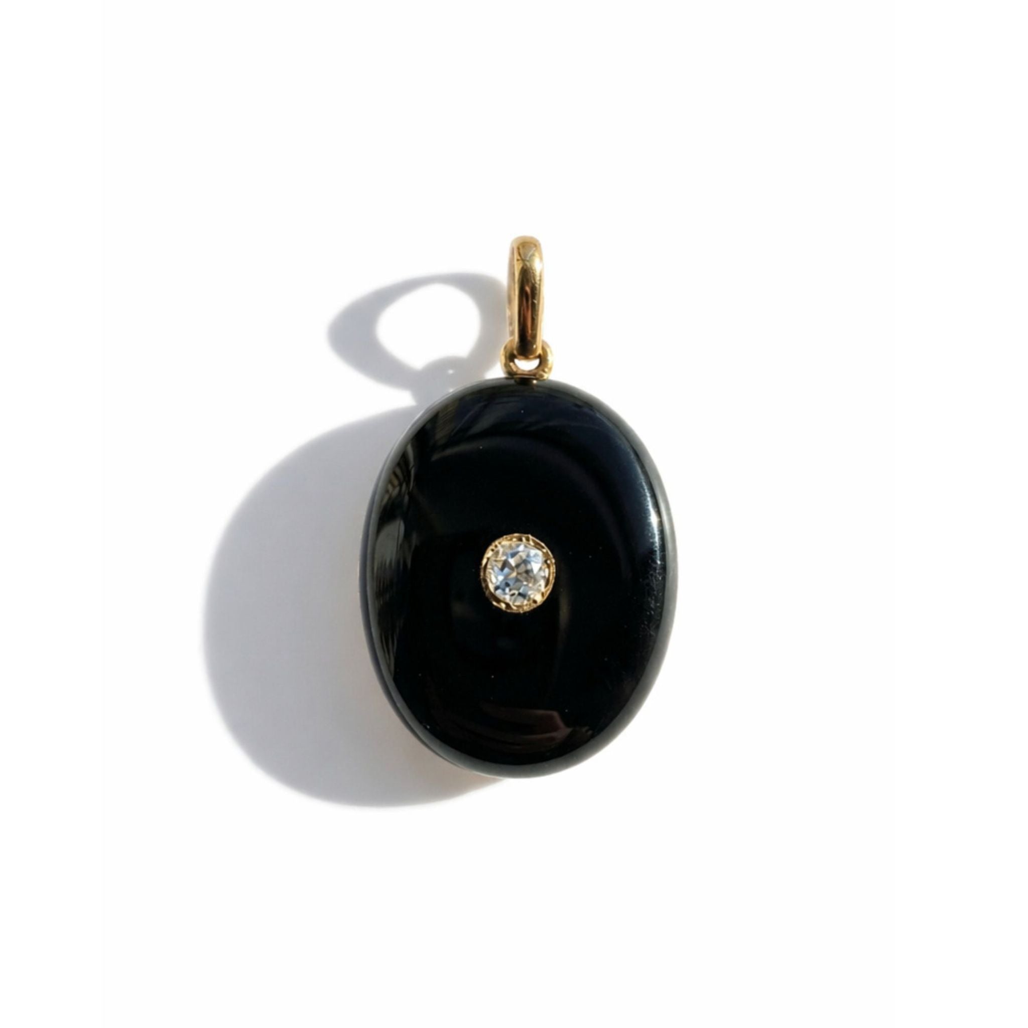 The Onyx and Diamond Locket features an elegant oval shape in sleek black onyx, accented by a small central diamond and set with a gold bail. Photographed on a white background with a soft shadow, this sophisticated piece embodies timeless elegance.
