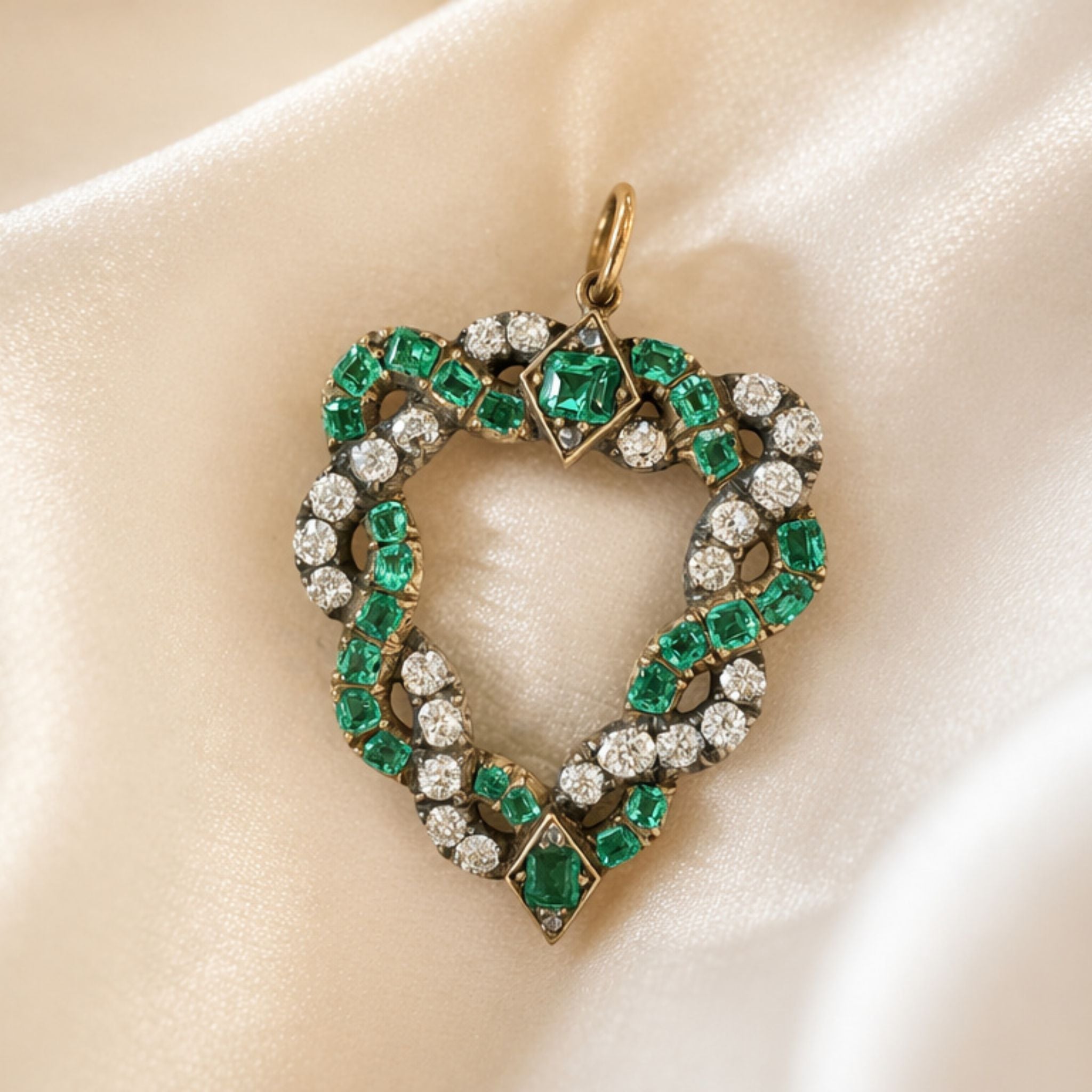 The Antique Emerald and Diamond Pendant, inspired by a vintage heart motif, showcases alternating cushion-shaped emeralds and shimmering old-cut diamonds set in gold, elegantly displayed atop soft, cream-colored satin fabric.