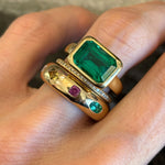 A hand wearing three gold rings: one is the East-West Bezel Set Emerald Cut Emerald Engagement Ring in Yellow Gold, another is adorned with small clear stones, and the third features various colorful gemstones arranged in a row.