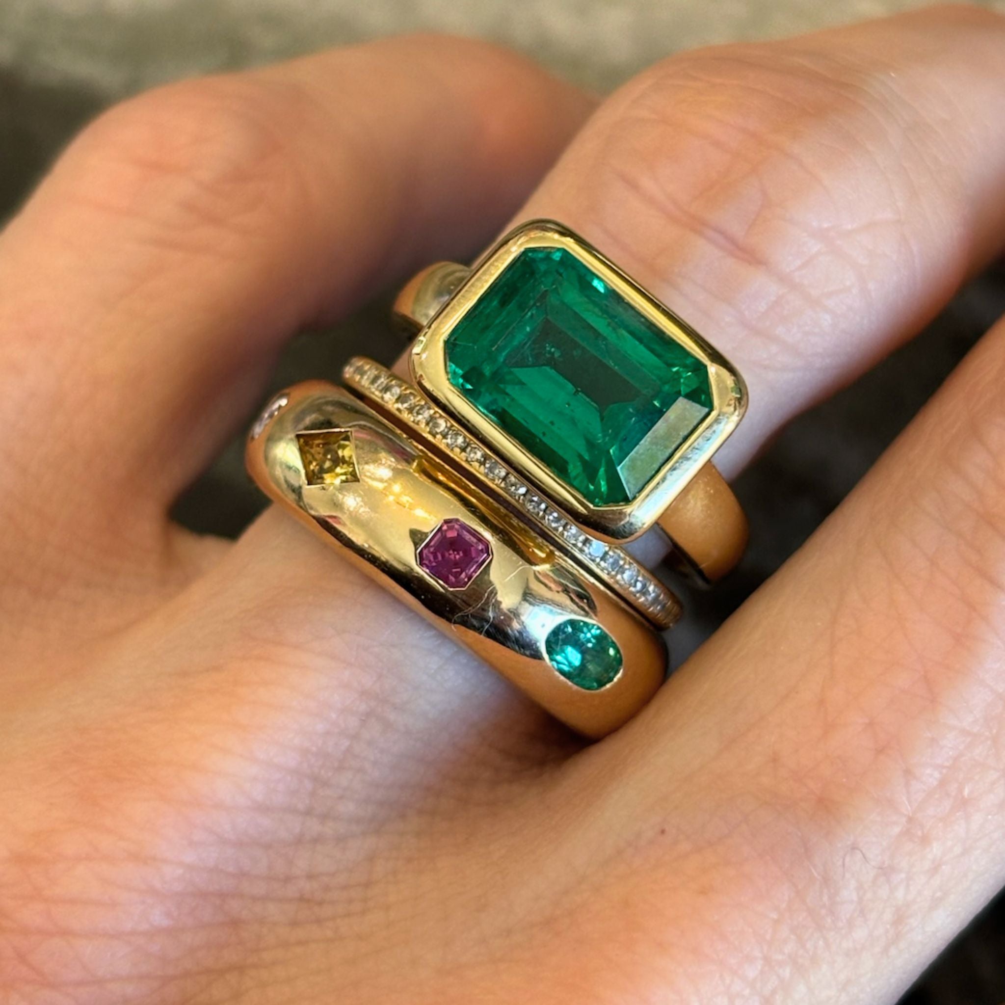 A hand wearing three gold rings: one is the East-West Bezel Set Emerald Cut Emerald Engagement Ring in Yellow Gold, another is adorned with small clear stones, and the third features various colorful gemstones arranged in a row.