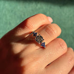 A hand wearing the Round Diamond Engagement Ring in White Gold, which features a large round center diamond flanked by kite-shaped blue sapphire side stones, set against a green textured background.