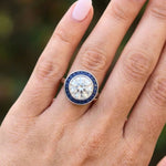 A hand with light pink nail polish wears the Round Brilliant Diamond with Channel Set Sapphire Halo Ring on the ring finger.