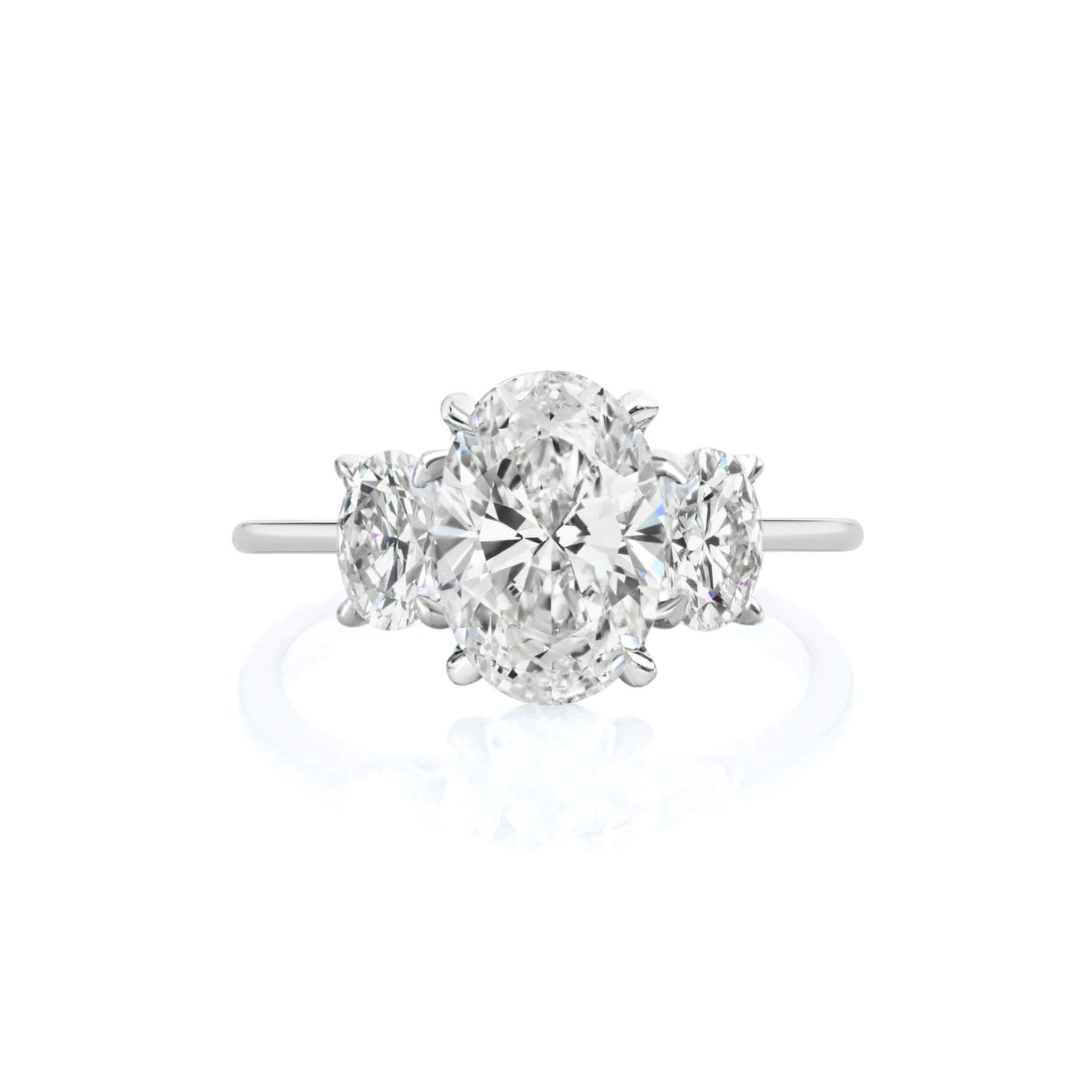 Introducing the Oval Diamond Engagement Ring with Oval Side Stones: a sophisticated engagement ring showcasing an oval-cut center diamond flanked by two smaller oval side stones, gracefully set on a slender white metal band against a pristine white background. This exquisite ring is the perfect choice to celebrate and honor life's most cherished moments.
