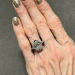 A hand adorned with gold glittery nail polish wears the Oval Shape Diamond Engagement Ring with Bullet Side Stones, featuring a large oval diamond on a platinum band accented by elegant tapered baguette side stones, showcased against a dark background.