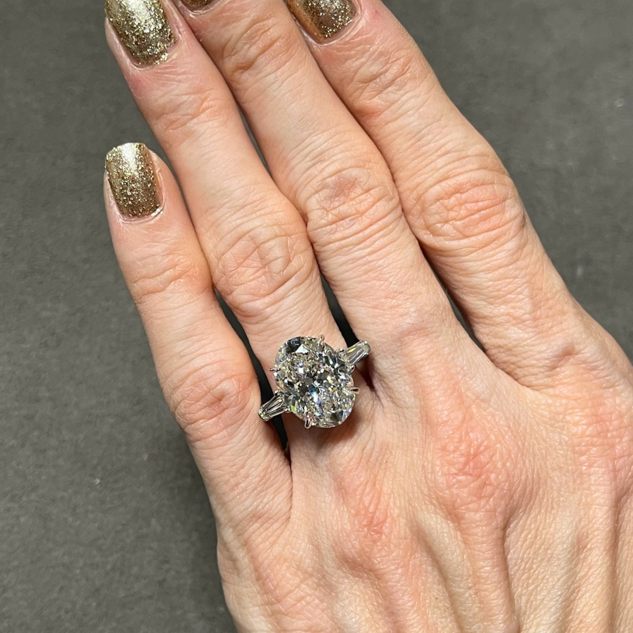 A hand adorned with gold glittery nail polish wears the Oval Shape Diamond Engagement Ring with Bullet Side Stones, featuring a large oval diamond on a platinum band accented by elegant tapered baguette side stones, showcased against a dark background.