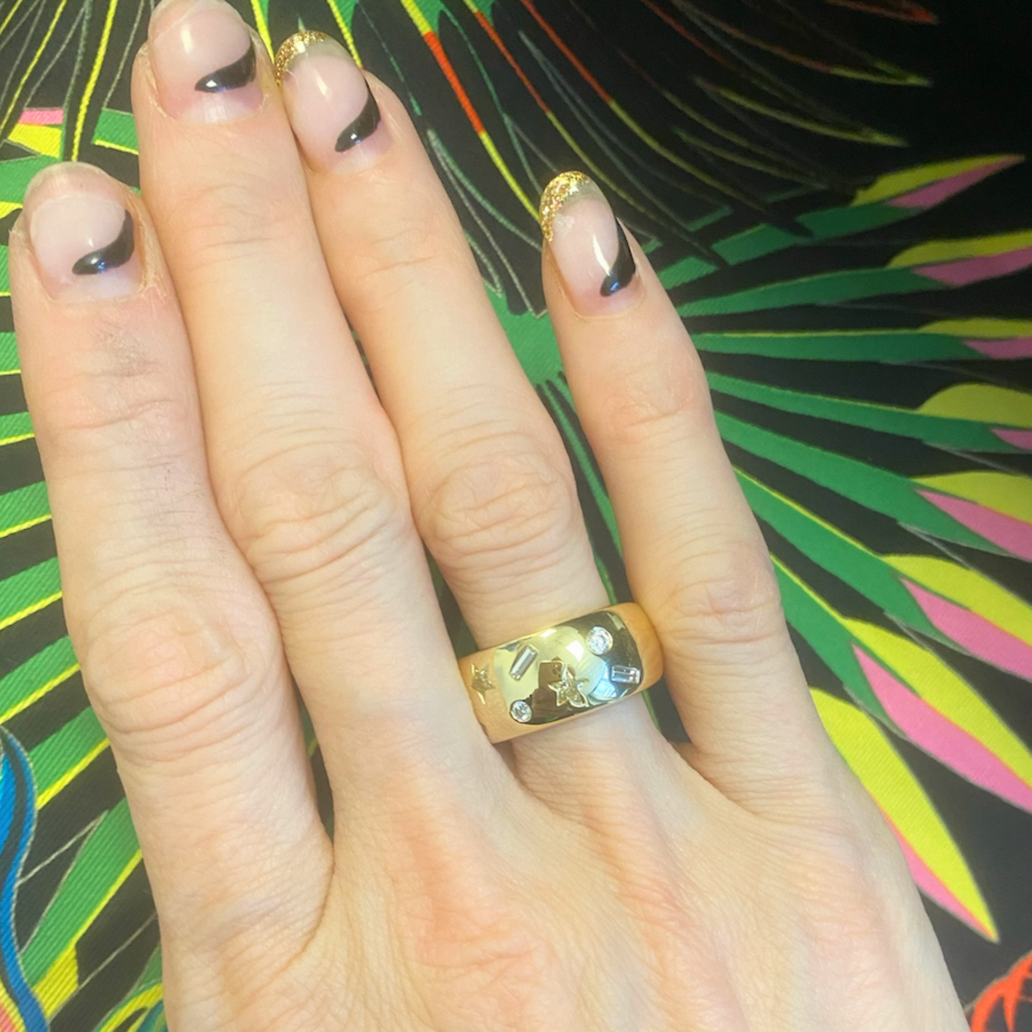 A hand with a black and gold-tipped French manicure rests on colorful, leafy fabric. The wide Yellow Diamond Celestial Cigar Band on the middle finger is crafted from 18K yellow gold with gemstones and star engravings.