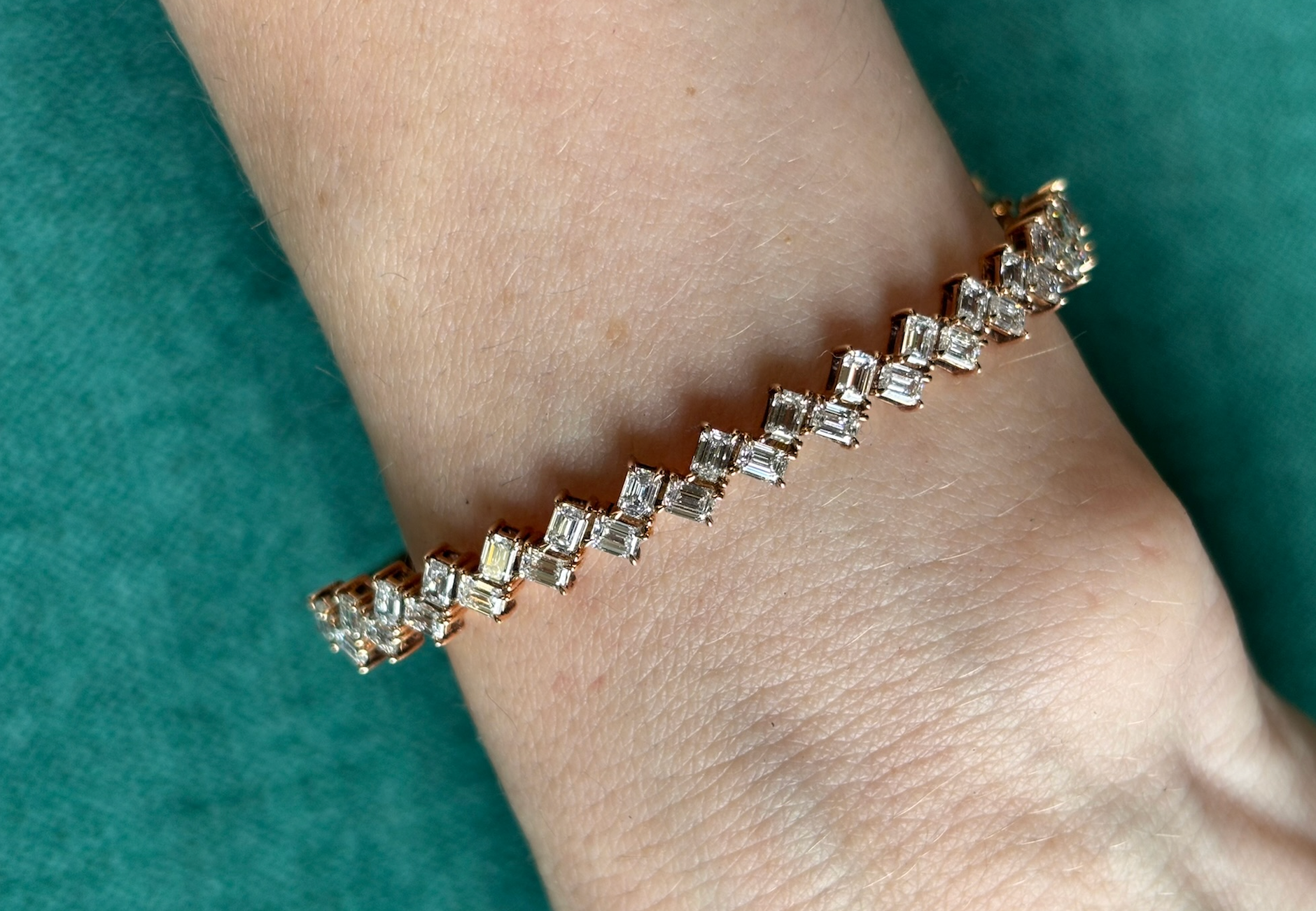 A close-up of a wrist wearing the Zig-Zag Baguette Diamond Bangle in Rose Gold, featuring sparkling square and rectangular-cut baguette diamonds, set against a green background.