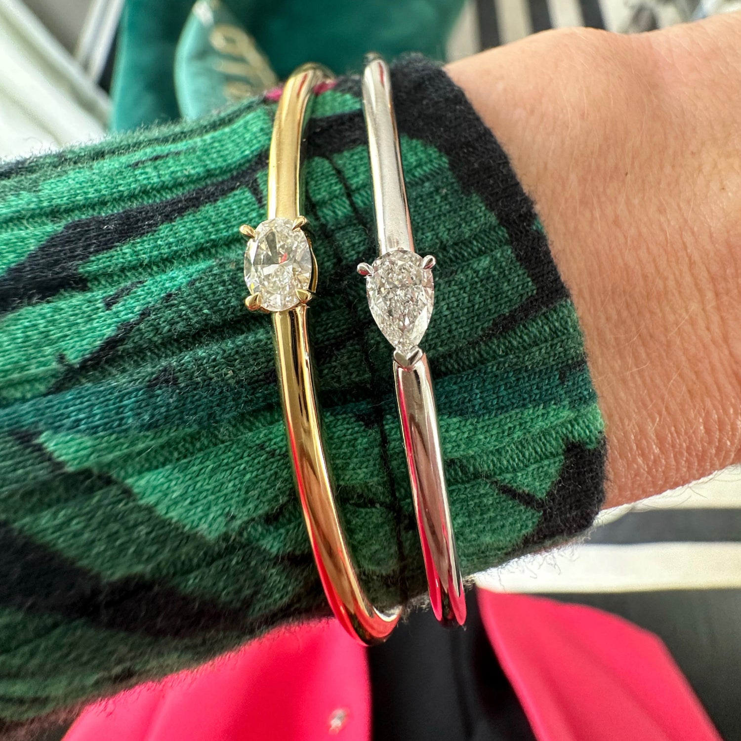 A close-up of a wrist wearing the East-West Oval Solitaire Diamond Bangle in 18K yellow gold and silver, paired with a green and black patterned sweater and a hint of pink garment underneath.
