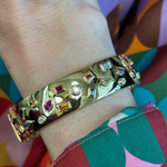 The Bezel Set Multi Shape Mixed Stone Bangle in Yellow Gold sparkles on the wrist, perfectly complementing a vibrant shirt with pink, green, orange, and blue patterns.