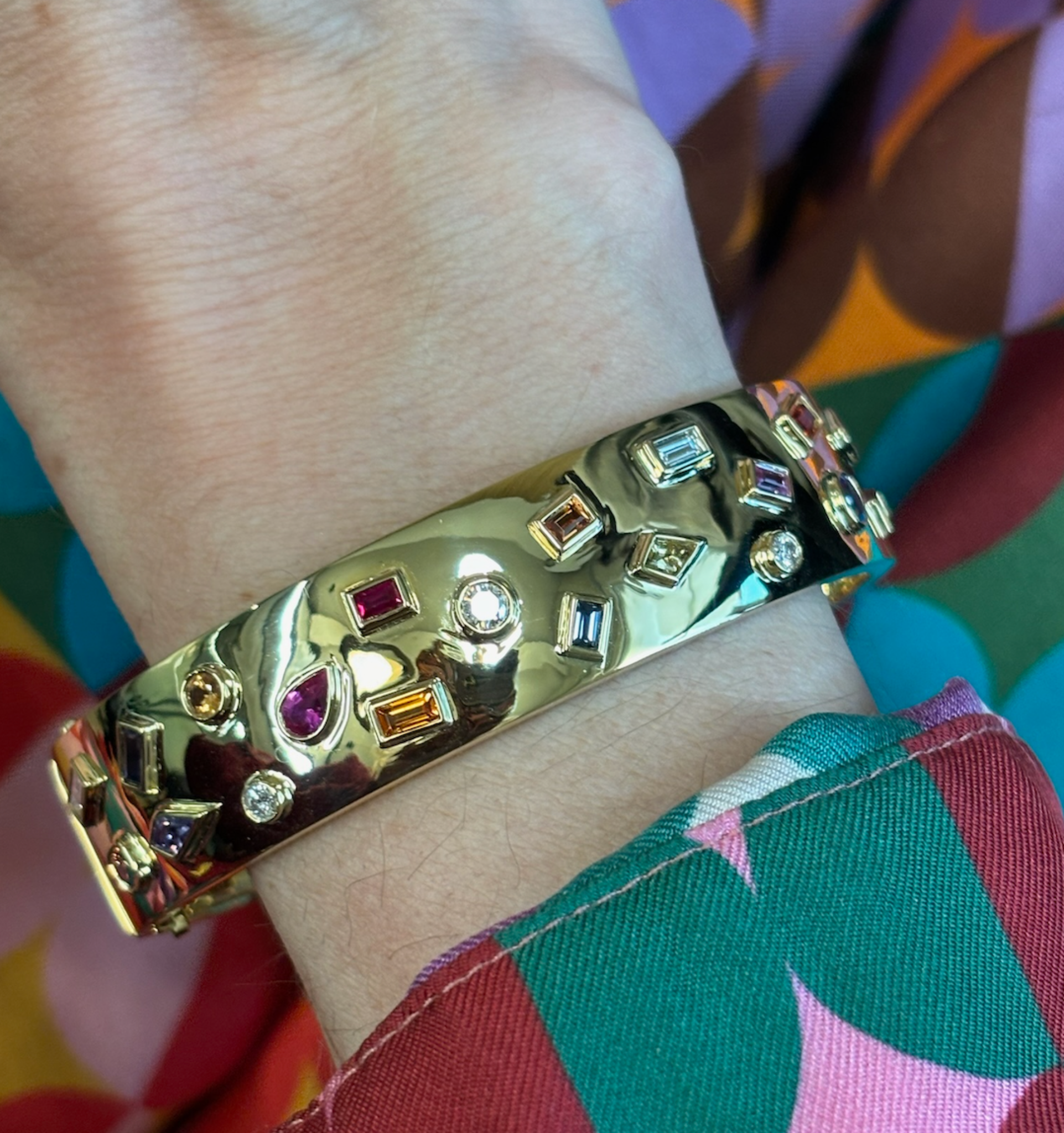 The Bespoke Bezel Set Multi Shape Mixed Stone Bangle in Yellow Gold, adorned with diamonds, shines on a wrist clad in vibrant geometric-patterned fabric of green, pink, orange, and blue hues.