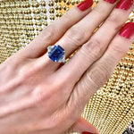 A hand with red-painted nails wears an Emerald Cut Sapphire Engagement Ring with Half Moon Diamond Side Stones set in silver, with gold bead strands blurred in the background.
