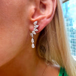 Close-up of a person’s ear with straight blonde hair, wearing a green top and Crescent Waterfall Earrings in white gold. The earrings feature pear diamonds extending downwards in a sparkling, leaf-inspired design.