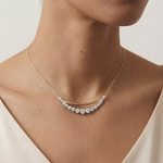 A woman wears the Coil Bar Pendant with Graduated Round Diamonds in Yellow Gold and Platinum, featuring a curved bar pendant adorned with sparkling diamonds, shown above a cream-colored top.