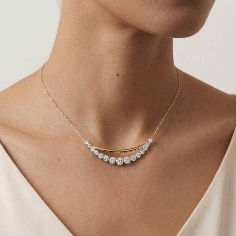A woman wears the Coil Bar Pendant with Graduated Round Diamonds in Yellow Gold and Platinum, featuring a curved bar pendant adorned with sparkling diamonds, shown above a cream-colored top.