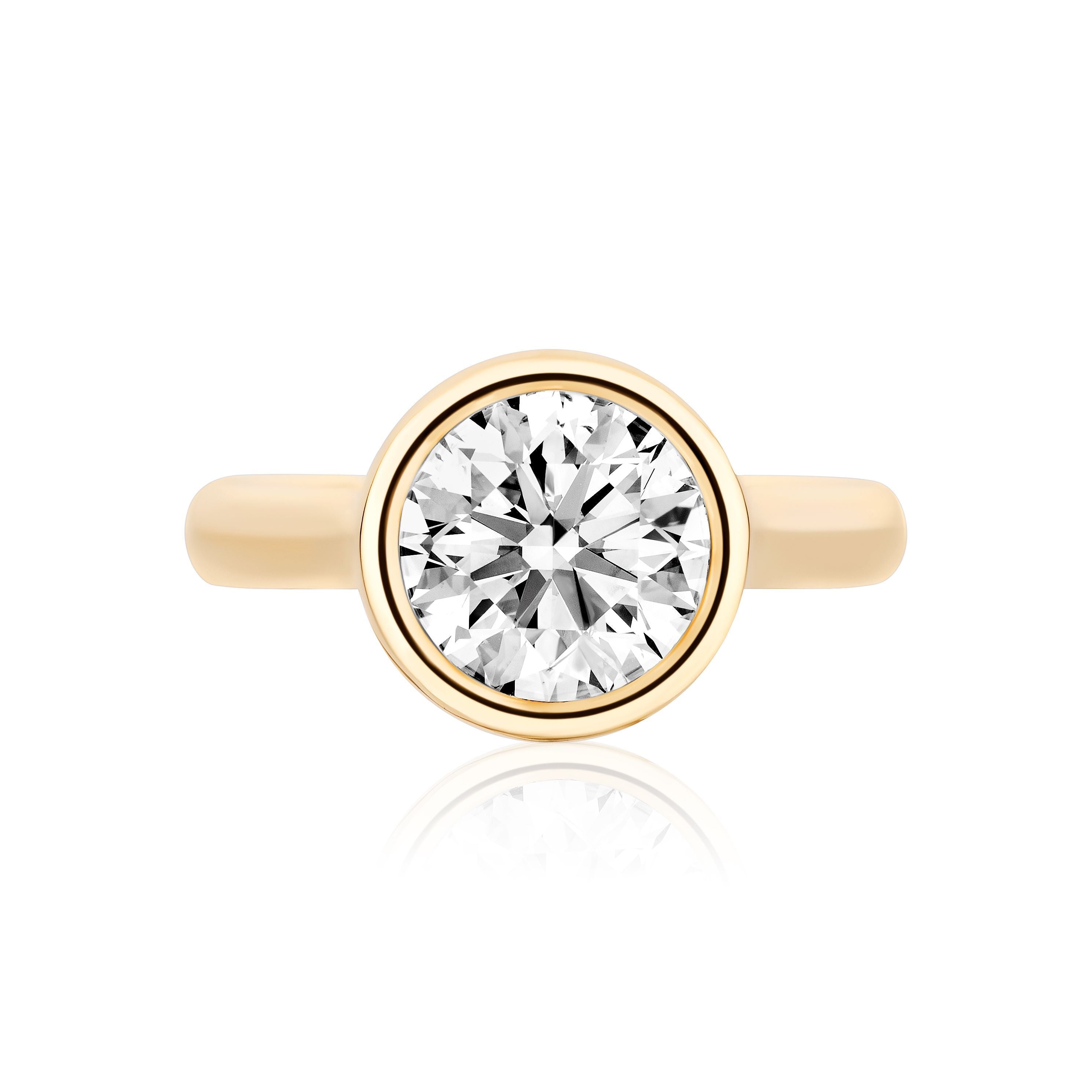 The Bezel Set Round Diamond Engagement Ring features a brilliant-cut round diamond at the center, showcased on a gold band against a white background with a subtle reflection.