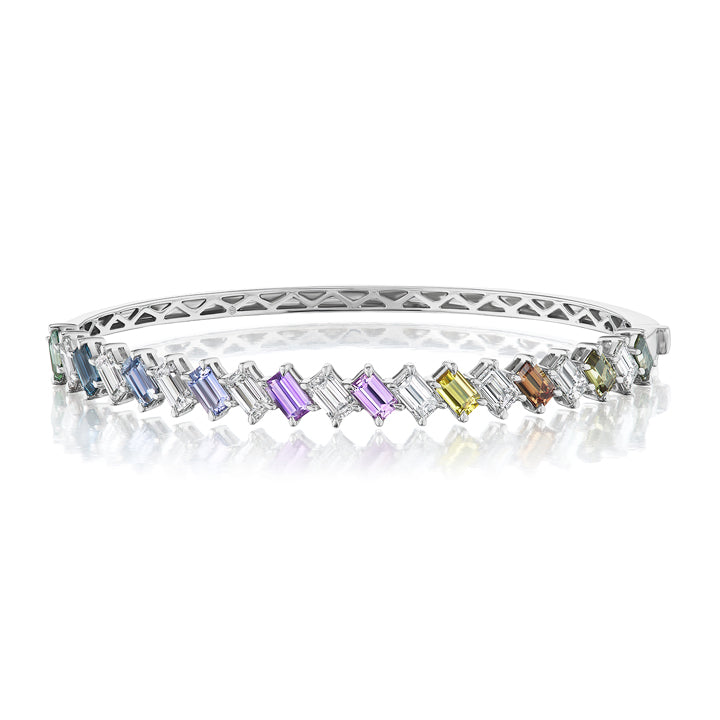 The Marching Emerald Cut Diamond and Multicolor Sapphire Bangle in White Gold features a row of vibrant sapphires and diamonds, elegantly set in a geometric pattern and displayed on a white reflective surface.