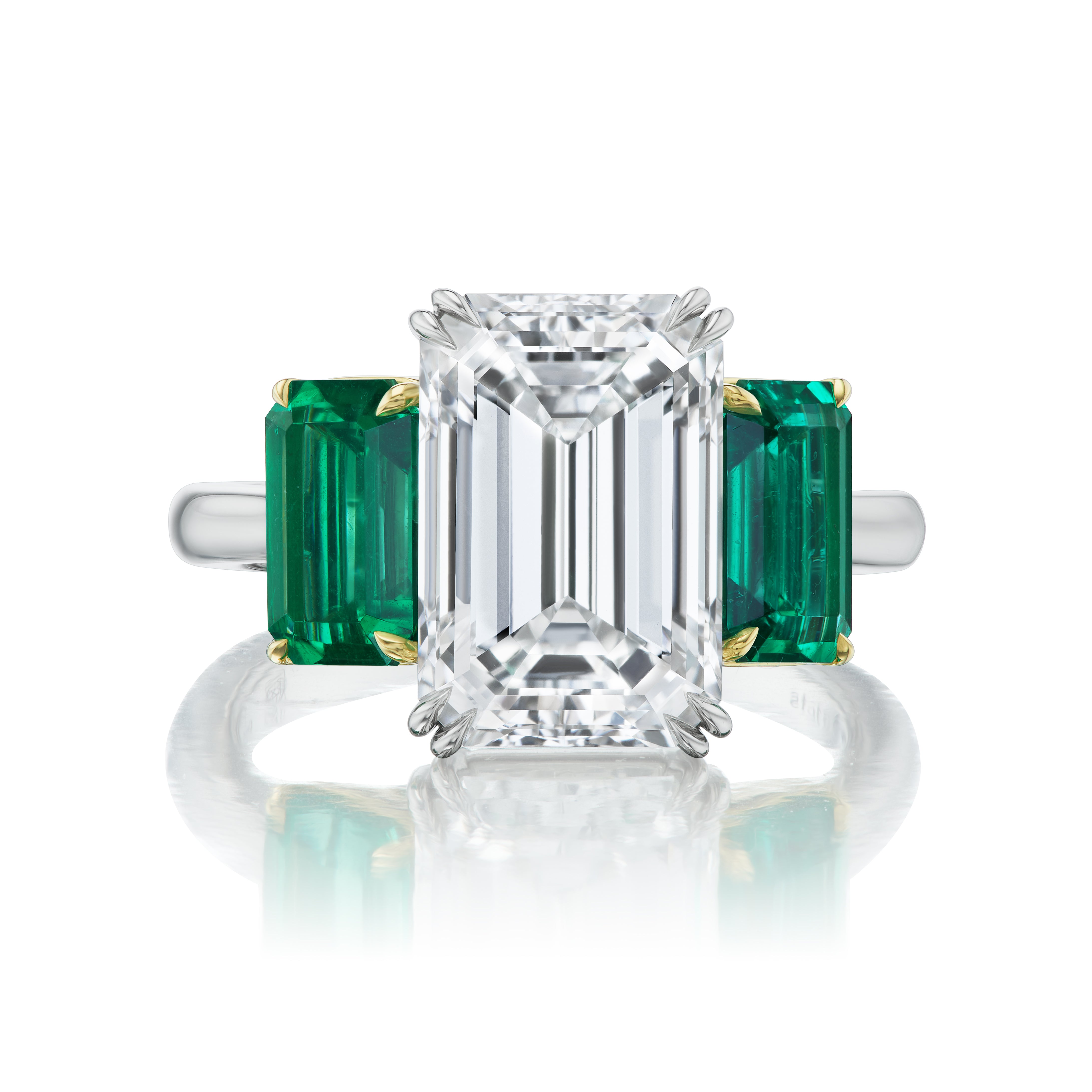 The Emerald Cut Diamond Engagement Ring features an emerald cut diamond center with emerald cut emerald side stones, set in 18K white gold prongs for the diamond and gold prongs for the emeralds.