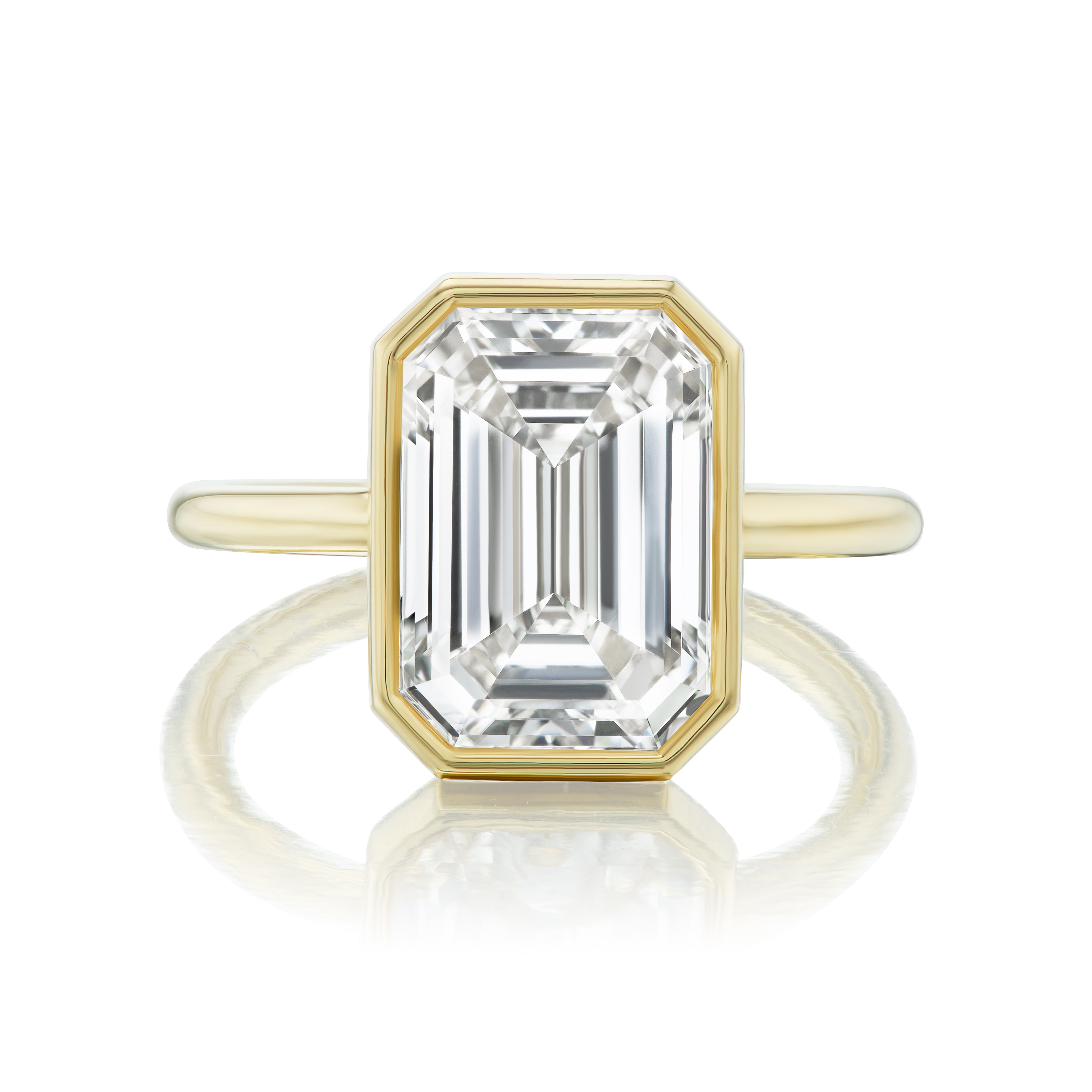 The Bezel Set Emerald Cut Diamond Engagement Ring features a rectangular, emerald-cut diamond in a sleek bezel setting atop a polished 18K yellow gold band.