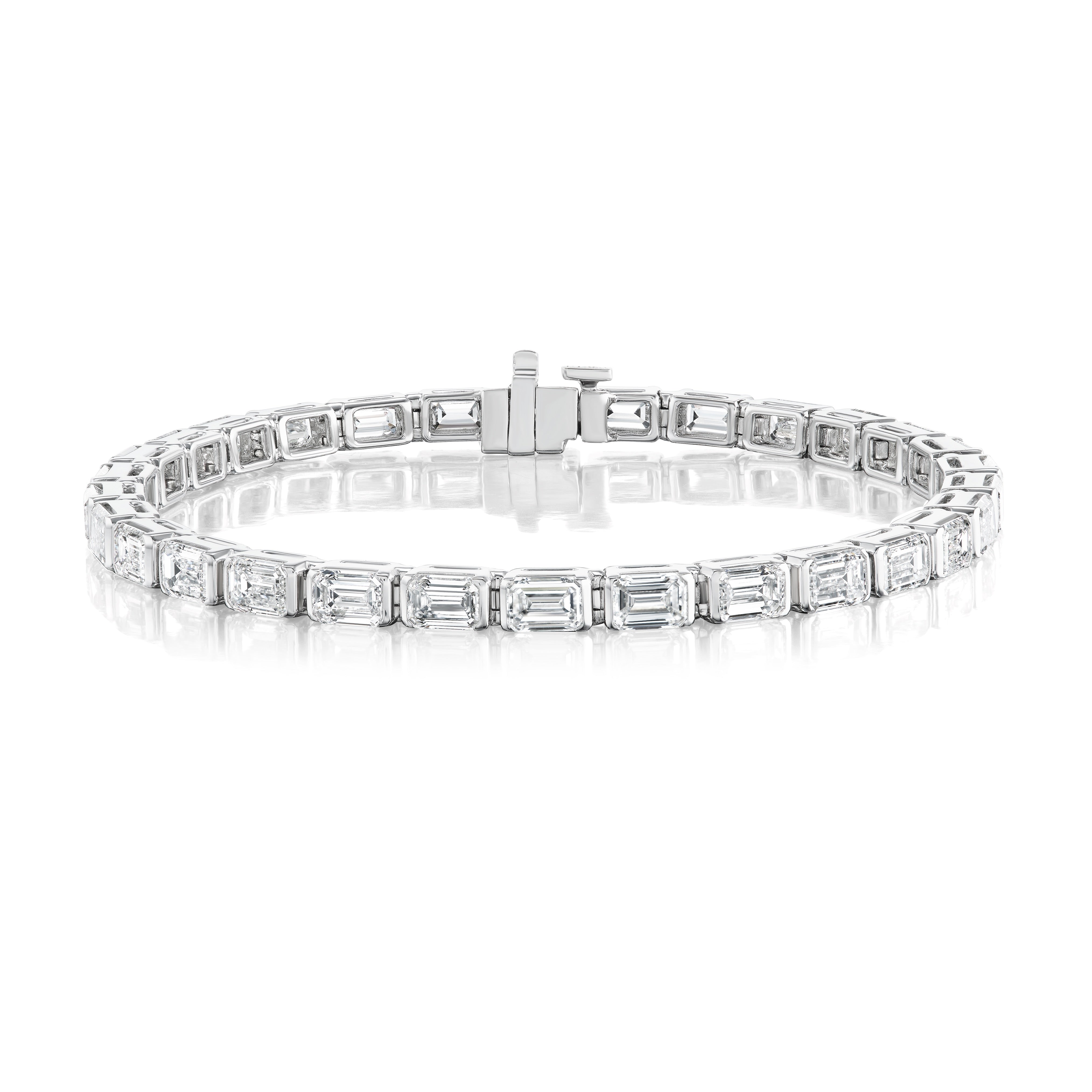 The East-West Half Bezel Set Emerald Cut Diamond Tennis Bracelet in Platinum showcases a row of emerald cut diamonds in individual half-bezel settings, beautifully displayed on a reflective white surface.