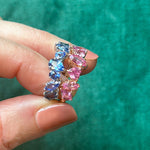 A hand holds two 18K gold rings against a textured green background: one is the Sapphire Mixed Shape Cluster Ring with blue sapphires, and the other features pink gemstones.