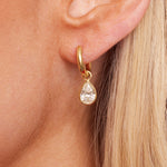 Close-up of a light-skinned person with straight blonde hair wearing Pear Diamond Hoops—18k yellow gold earrings set with SI clarity diamonds and a teardrop-shaped, clear gemstone pendant.