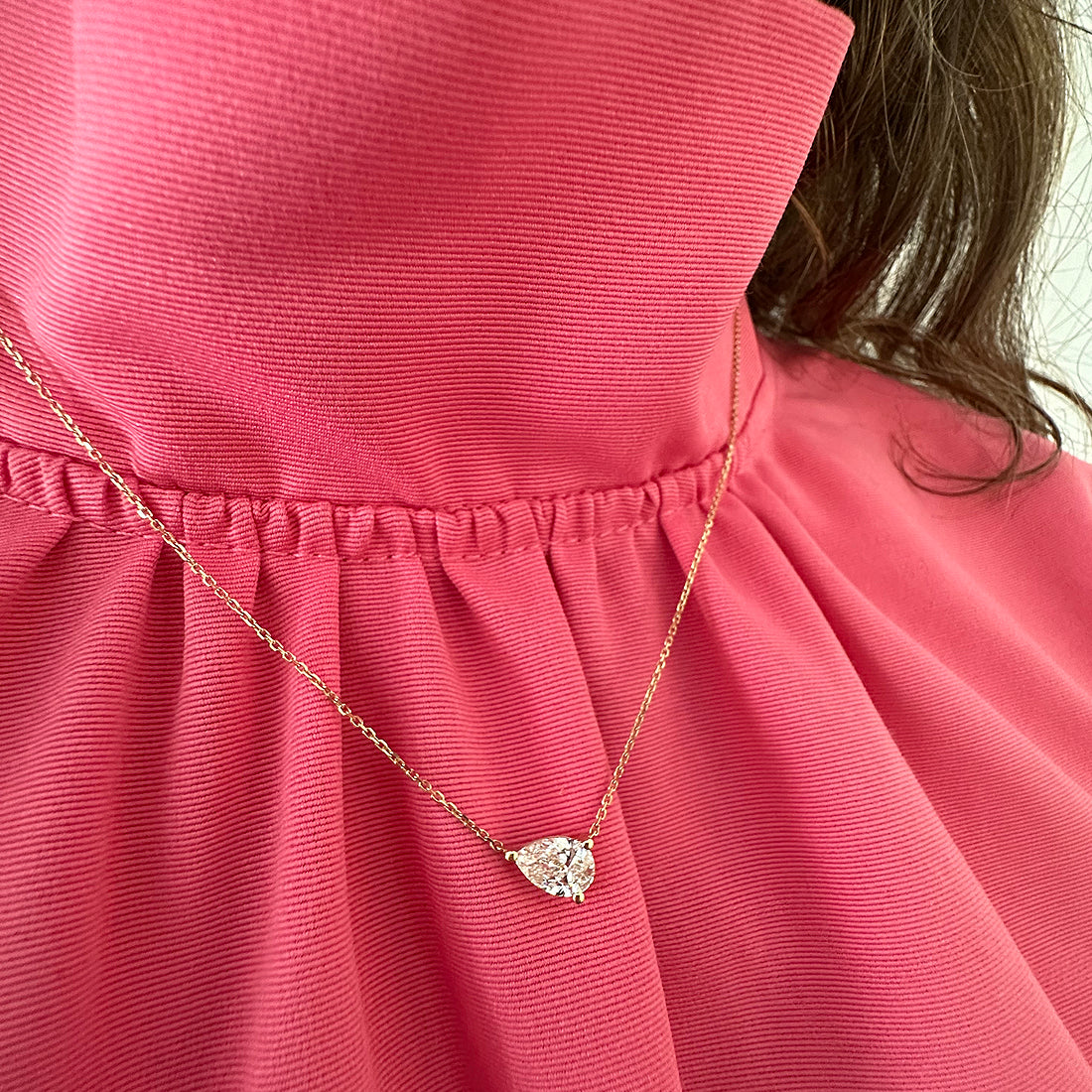 A close-up of the East-West Set Pear Pendant Necklace in 18K yellow gold, featuring a teardrop gemstone, worn over a pink textured high-collar blouse. Brown hair appears partially on the right.