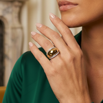 A woman in a green blouse rests her hand on her face, showcasing the Coil Dome Ring with Pavé Diamond Borders in Yellow Gold. A blurred marble fireplace appears in the background.